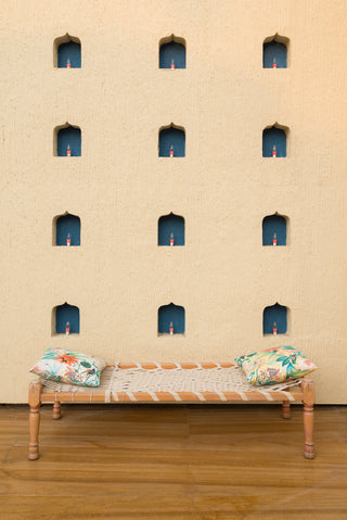Wooden Cot with flower cushions against a beige wall with small windows.